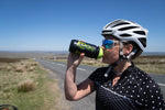 A female cyclists stood by the side of a road over a moorland drinking from a black and green Voom bottle