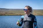 A woman in a wetsuit stood by a lake holding a half eaten Voom electro energy bar with a smile on her face.